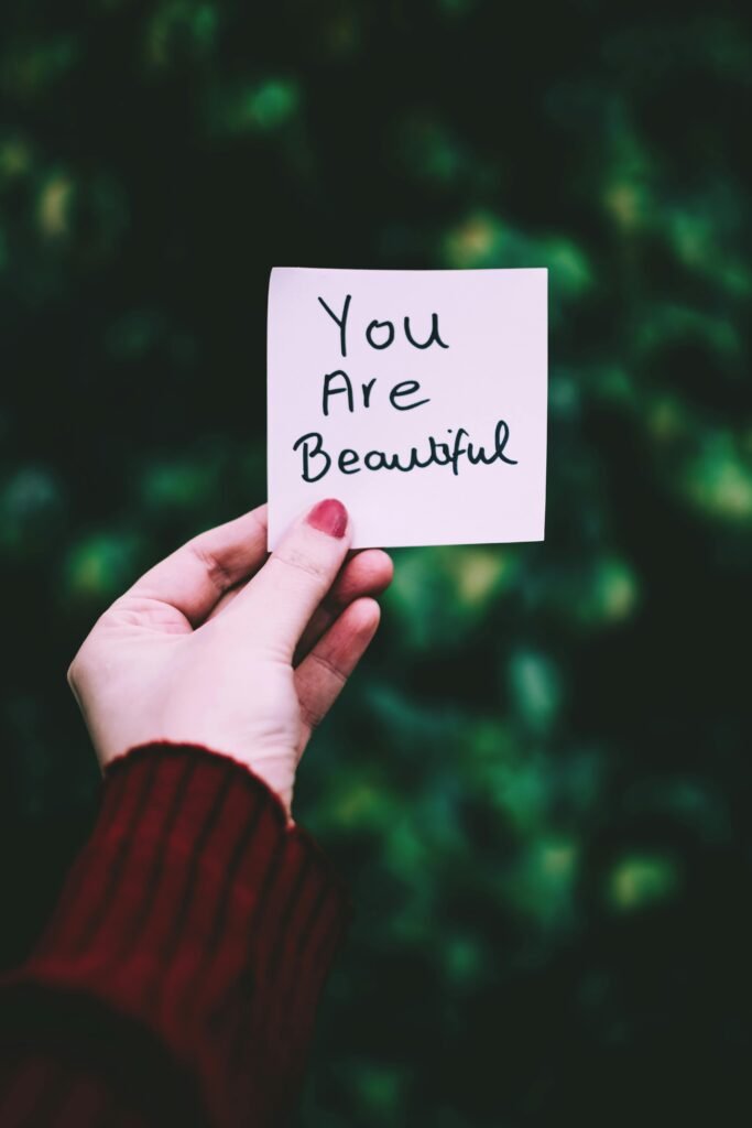 A close-up of a hand holding a 'You Are Beautiful' note against a blurred green background.