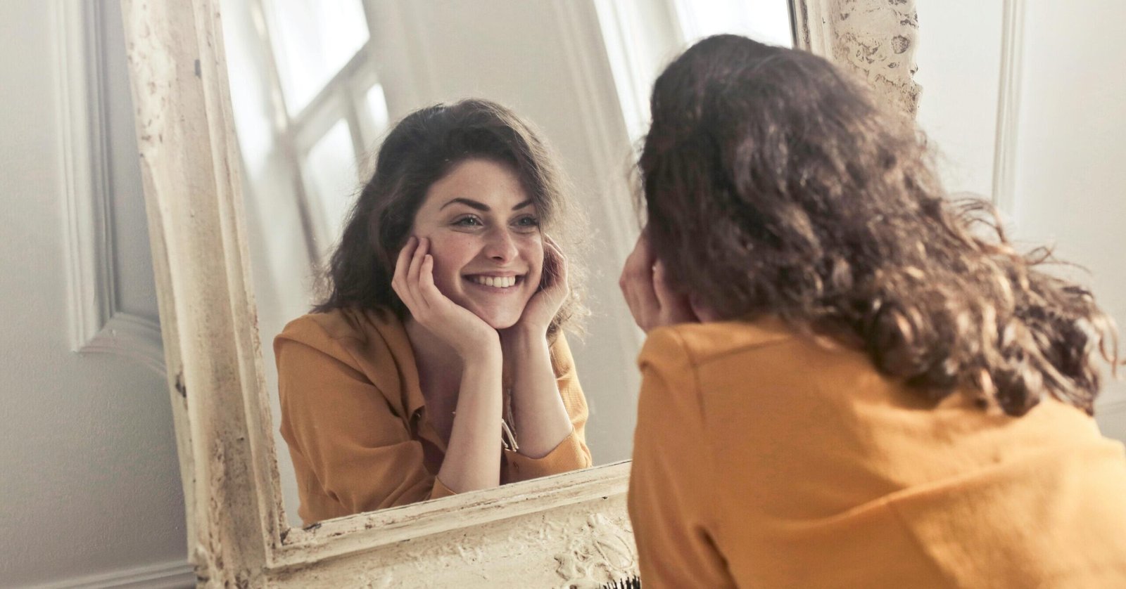 A cheerful woman smiles at her reflection in a vintage-style mirror, exuding positivity and warmth.